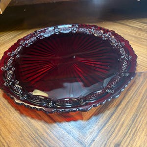 May include: A deep red, oval glass serving dish. The dish has a ribbed interior and a decorative border with a repeating pattern of floral and grape clusters. The dish is likely vintage and suitable for serving food.