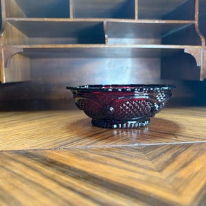 May include: A ruby red glass bowl with a decorative pattern. The bowl has a scalloped rim and a pedestal base. The bowl is sitting on a wooden surface with a geometric pattern. The background includes a wooden cabinet.