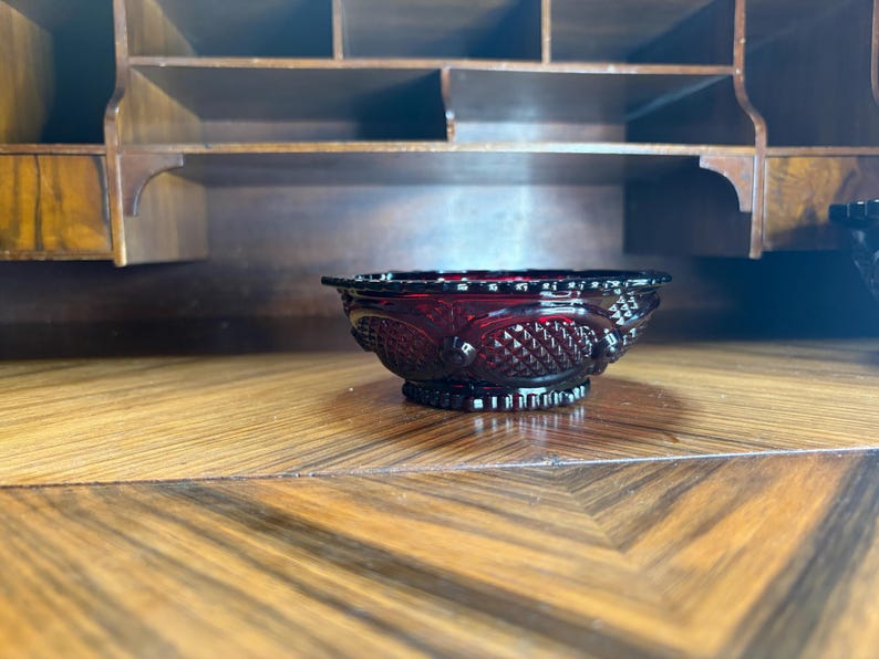 May include: A ruby red glass bowl with a decorative pattern. The bowl has a scalloped rim and a pedestal base. The bowl is sitting on a wooden surface with a geometric pattern. The background includes a wooden cabinet.