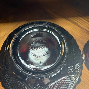 May include: Black glass bowl with a diamond and textured pattern. The bottom of the bowl has the text "AVON 1886-1986". The bowl has a scalloped edge and is sitting on a wooden surface.