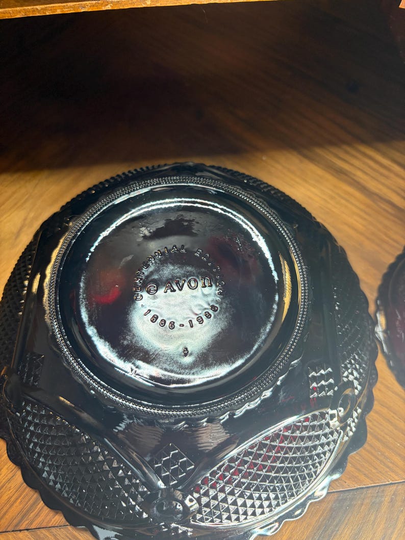 May include: Black glass bowl with a diamond and textured pattern. The bottom of the bowl has the text "AVON 1886-1986". The bowl has a scalloped edge and is sitting on a wooden surface.
