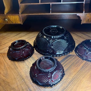 May include: Four ruby red glass bowls with a textured pattern are arranged on a wooden surface. The bowls vary in size, with a larger bowl in the center and smaller bowls surrounding it. The bowls have a decorative design.