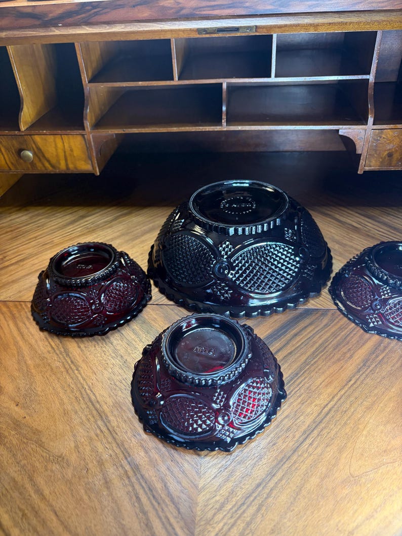 May include: Four ruby red glass bowls with a textured pattern are arranged on a wooden surface. The bowls vary in size, with a larger bowl in the center and smaller bowls surrounding it. The bowls have a decorative design.