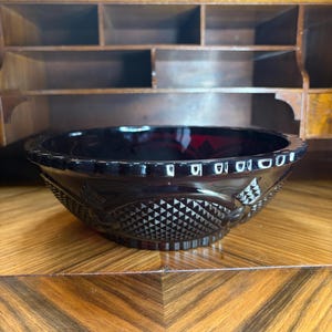 May include: A dark red glass bowl with a decorative diamond and floral pattern. The bowl has a scalloped rim and sits on a wooden surface. The bowl is likely vintage or antique.