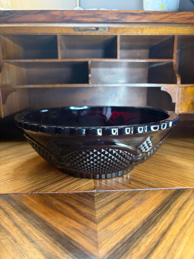 May include: A dark red glass bowl with a decorative diamond and floral pattern. The bowl has a scalloped rim and sits on a wooden surface. The bowl is likely vintage or antique.