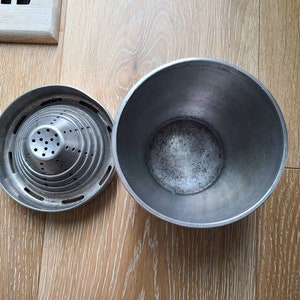 Puede incluir: Un exprimidor de c&iacute;tricos de metal plateado con un colador y un taz&oacute;n separado. El exprimidor tiene una forma c&oacute;nica con m&uacute;ltiples capas de agujeros. El taz&oacute;n es redondo y profundo.