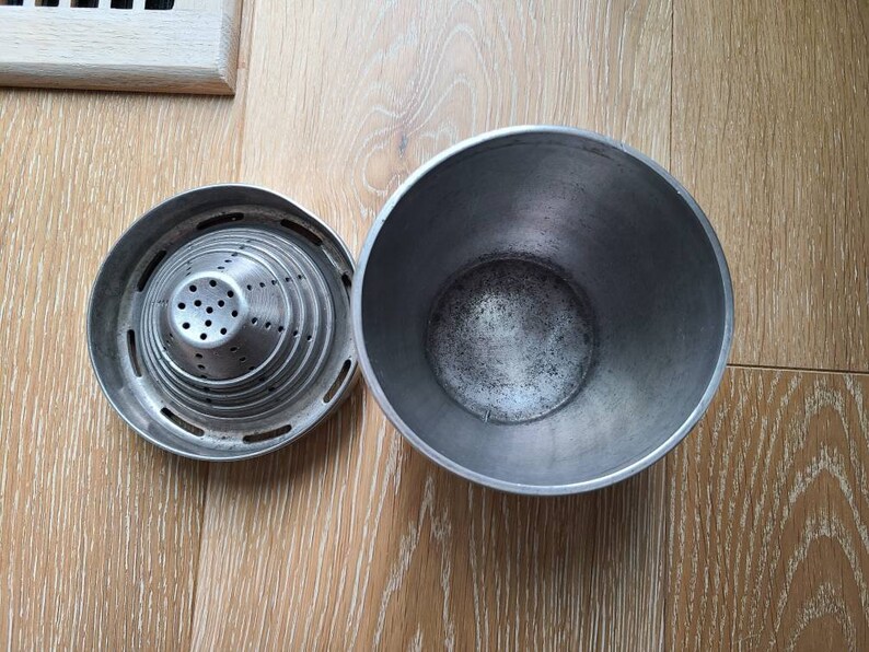 Puede incluir: Un exprimidor de c&iacute;tricos de metal plateado con un colador y un taz&oacute;n separado. El exprimidor tiene una forma c&oacute;nica con m&uacute;ltiples capas de agujeros. El taz&oacute;n es redondo y profundo.