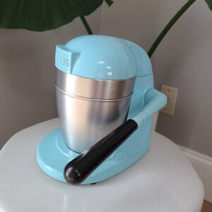 Puede incluir: Un exprimidor de c&iacute;tricos el&eacute;ctrico azul claro con un recipiente de metal plateado y un mango negro. El exprimidor tiene la palabra "KING" impresa en el lateral.