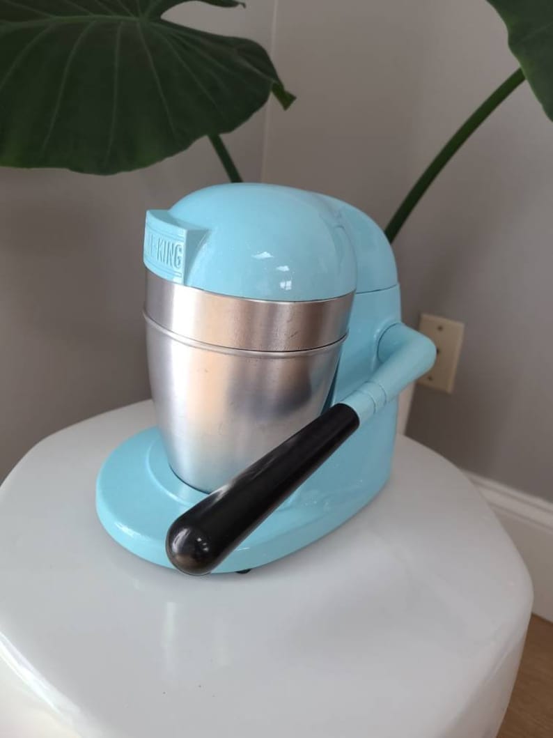 Puede incluir: Un exprimidor de c&iacute;tricos el&eacute;ctrico azul claro con un recipiente de metal plateado y un mango negro. El exprimidor tiene la palabra "KING" impresa en el lateral.