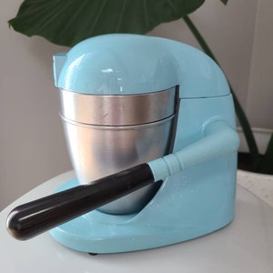 Puede incluir: Un exprimidor de c&iacute;tricos el&eacute;ctrico azul claro con un mango negro y un recipiente de acero inoxidable. El exprimidor est&aacute; sobre una superficie blanca.