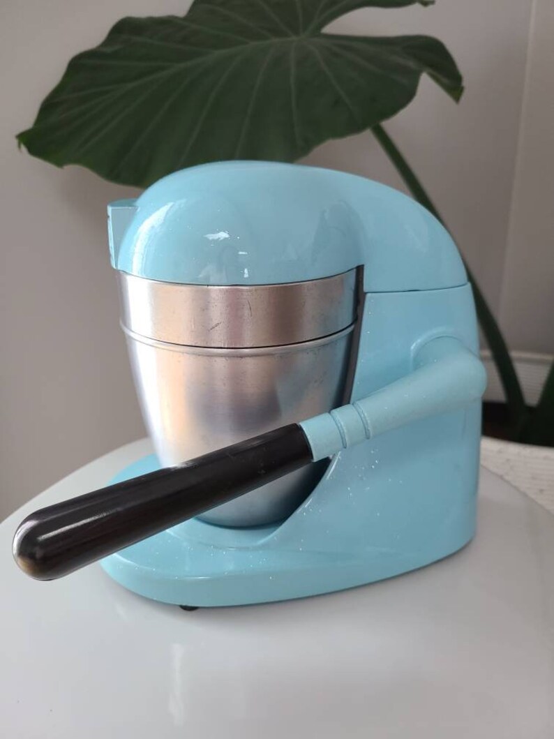 Puede incluir: Un exprimidor de c&iacute;tricos el&eacute;ctrico azul claro con un mango negro y un recipiente de acero inoxidable. El exprimidor est&aacute; sobre una superficie blanca.