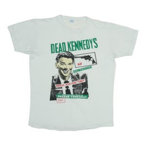 May include: White t-shirt featuring the text "DEAD KENNEDYS" in green above a graphic of a man with a gun and the words "KILL THE POOR". Below the graphic is the text "SMILE AND SAY, DIE!" and "DEFEND YOURSELF!"