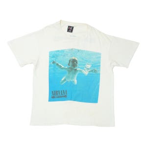 May include: A white short-sleeve t-shirt featuring a graphic print of a baby swimming underwater toward a dollar bill. The graphic includes the text NIRVANA and NEVERMIND in a dark font at the bottom. The shirt has a crew neckline and a brand tag.