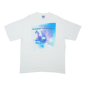 May include: A white short-sleeved t-shirt featuring a graphic print on the chest. The graphic displays a blue-toned image of a person wearing a hat, with the text "tim mcgraw" and "a place in the sun" printed in small dark letters above the figure.