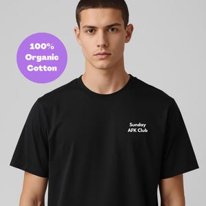 Op de afbeelding: Een zwart T-shirt met ronde hals en de tekst "Sunday AFK Club" in het wit op de borst. Een paarse cirkel met de tekst "100% Organic Cotton" staat in de linkerbovenhoek. Het T-shirt wordt door een persoon gedragen.