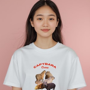 Könnte beinhalten: Weißes T-Shirt mit einer Grafik von fünf Capybaras und dem Text "CAPYBARA Crew" in Rot. Die Capybaras sind in verschiedenen Braun- und Beigetönen gehalten. Das T-Shirt wird von einer Person mit langen dunklen Haaren getragen.