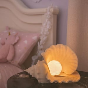 Puede incluir: Lámpara con forma de concha marina, emitiendo una luz cálida. La lámpara blanquecina está sobre una mesa redonda oscura. En el fondo, una almohada rosa con diseño de unicornio y un arcoíris decorativo.