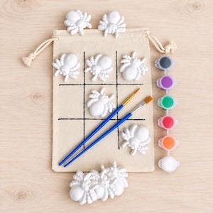 May include: A beige drawstring canvas bag printed with a black tic-tac-toe grid, accompanied by several white spider-shaped figurines, two blue-handled paintbrushes, and a strip of six small paint pots in black, purple, green, red, orange, and white.