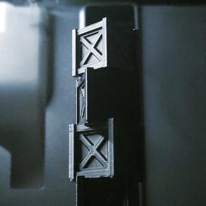 May include: Three small, dark grey, rectangular structures stacked vertically. Each has a square frame with an 'X' design in the centre. The structures are connected and appear to be 3D-printed.