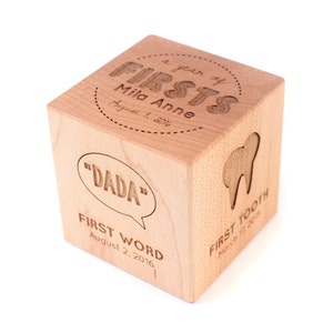 May include: A wooden cube with engraved text commemorating a child's first year. The text includes "a year of FIRSTS", "Mila Anne", "August 1, 2016", "DADA", "FIRST WORD", "August 2, 2016", "FIRST TOOTH", and "March 15, 2015".