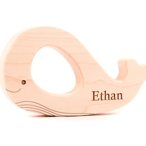 May include: A light-colored wooden whale-shaped baby teether. The teether has a circular opening and a small tail fin. The name "Ethan" is engraved on the side. The whale has a smiling face and curved lines.