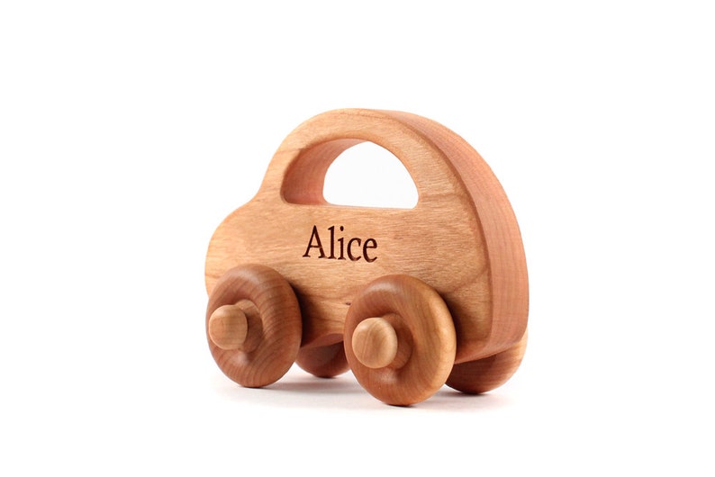 Pode incluir: Um carro de brinquedo de madeira com acabamento castanho claro. O carro tem uma carroceria arredondada com um recorte para uma al&ccedil;a e quatro rodas de madeira. O nome "Alice" est&aacute; gravado na lateral. Um brinquedo cl&aacute;ssico para crian&ccedil;as.
