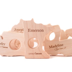 May include: Five wooden state-shaped teethers with personalized names, including Ryder, Emerson, Madeline, Joseph, and Lila. Each teether features the text "Locally Grown" and is designed for babies.