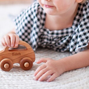 Pode incluir: Um carro de brinquedo de madeira com o nome "Edith" gravado na lateral. O carro &eacute; feito de madeira clara e tem quatro rodas de madeira. A crian&ccedil;a est&aacute; vestindo uma blusa xadrez preta e branca.