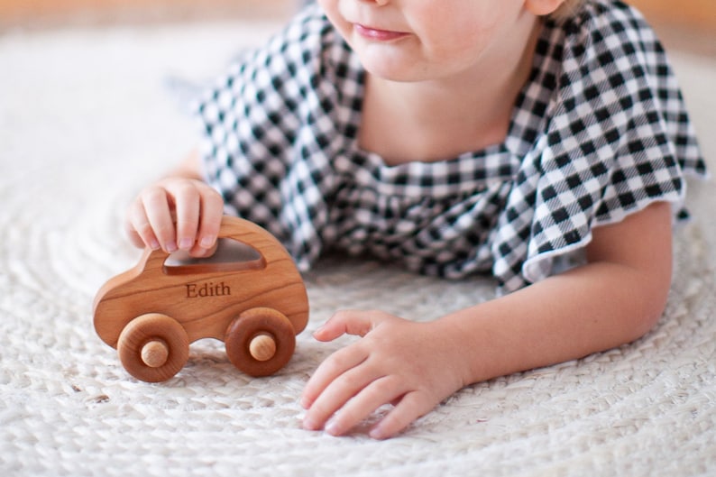 Pode incluir: Um carro de brinquedo de madeira com o nome "Edith" gravado na lateral. O carro &eacute; feito de madeira clara e tem quatro rodas de madeira. A crian&ccedil;a est&aacute; vestindo uma blusa xadrez preta e branca.