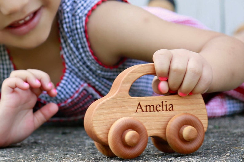 Pode incluir: Um carro de brinquedo de madeira com o nome "Amelia" gravado na lateral. O carro &eacute; feito de madeira clara e tem quatro rodas redondas. O carro est&aacute; sendo segurado por uma pessoa.