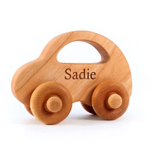 Pode incluir: Carro de brinquedo de madeira com quatro rodas e um teto curvo. O nome "Sadie" est&aacute; gravado na lateral. O carro &eacute; feito de madeira clara e tem um acabamento liso.