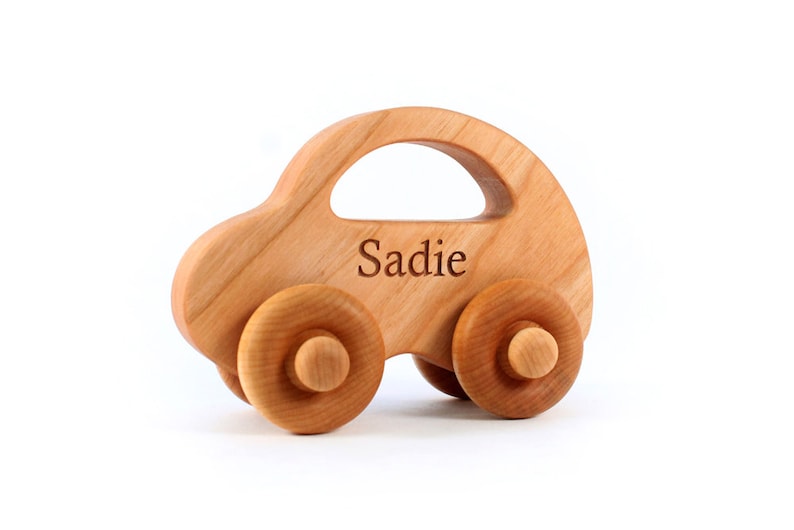 Pode incluir: Carro de brinquedo de madeira com quatro rodas e um teto curvo. O nome "Sadie" est&aacute; gravado na lateral. O carro &eacute; feito de madeira clara e tem um acabamento liso.