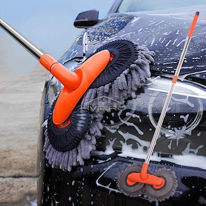 Puede incluir: Un cepillo para lavar coches con un mango metálico largo y extensible y un agarre naranja. El cabezal del cepillo tiene un diseño circular con una superficie de limpieza de microfibra gris. El cepillo se muestra limpiando un coche negro con agua jabonosa.