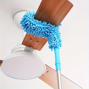 Könnte beinhalten: Ein blauer Mikrofaserduster reinigt einen Deckenventilatorflügel aus Holz. Der Duster hat einen langen, ausziehbaren Griff, um hohe Stellen zu erreichen. Der Ventilator ist weiß mit Holzflügeln.