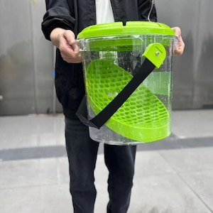 May include: A clear plastic salad spinner with a bright green lid and inner basket. The spinner has a black strap for carrying. The design includes a handle and a textured basket for washing and drying greens. The product is designed for kitchen use.