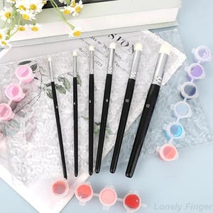 Könnte beinhalten: Ein Set von Malpinseln mit schwarzen Griffen und weißen Borsten, nummeriert. Kleine Farbtöpfe in Rosa, Rot, Blau und Lila sind ebenfalls zu sehen, zusammen mit einem Blumenarrangement.