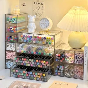 May include: A collection of clear acrylic desk organizers filled with various colored markers and pens. The modular storage units feature multiple drawers and compartments, neatly holding art supplies in a variety of vibrant hues and shades.