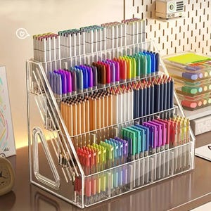 Peut inclure: Un organisateur de bureau à étages en acrylique transparent doté de quatre étagères inclinées remplies d'une grande collection de stylos, marqueurs et crayons colorés classés par couleur, avec des panneaux latéraux triangulaires.