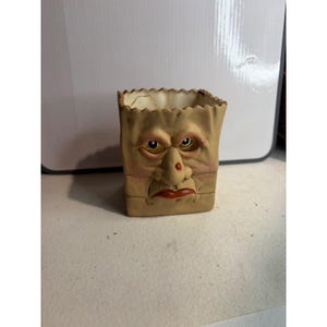 May include: A beige ceramic container shaped like a paper bag with a sculpted human face. The face features prominent eyes, a nose with a red spot, and red lips. The top edge of the container has a jagged, uneven rim resembling a crumpled bag.