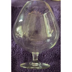 May include: A clear glass snifter with a wide, rounded bowl that tapers toward a narrow opening at the top. The vessel features a short, sturdy stem connected to a circular base, designed for holding spirits or as a decorative glass piece.