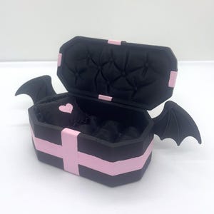 以下が含まれることがあります： ピンクのアクセントとコウモリの羽が付いた、黒い棺型の箱。箱は開いており、ピンクのハートが付いた黒い内側が見えます。棺はピンクのストライプとアクセントで装飾されています。