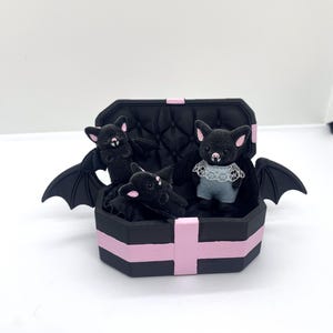 以下が含まれることがあります： ピンクのアクセントが付いた黒い棺型の箱に、3匹のぬいぐるみコウモリが入っています。2匹のコウモリは翼を広げ、3匹目は白いレースの襟が付いた青い服を着ています。コウモリはピンクの耳をしています。