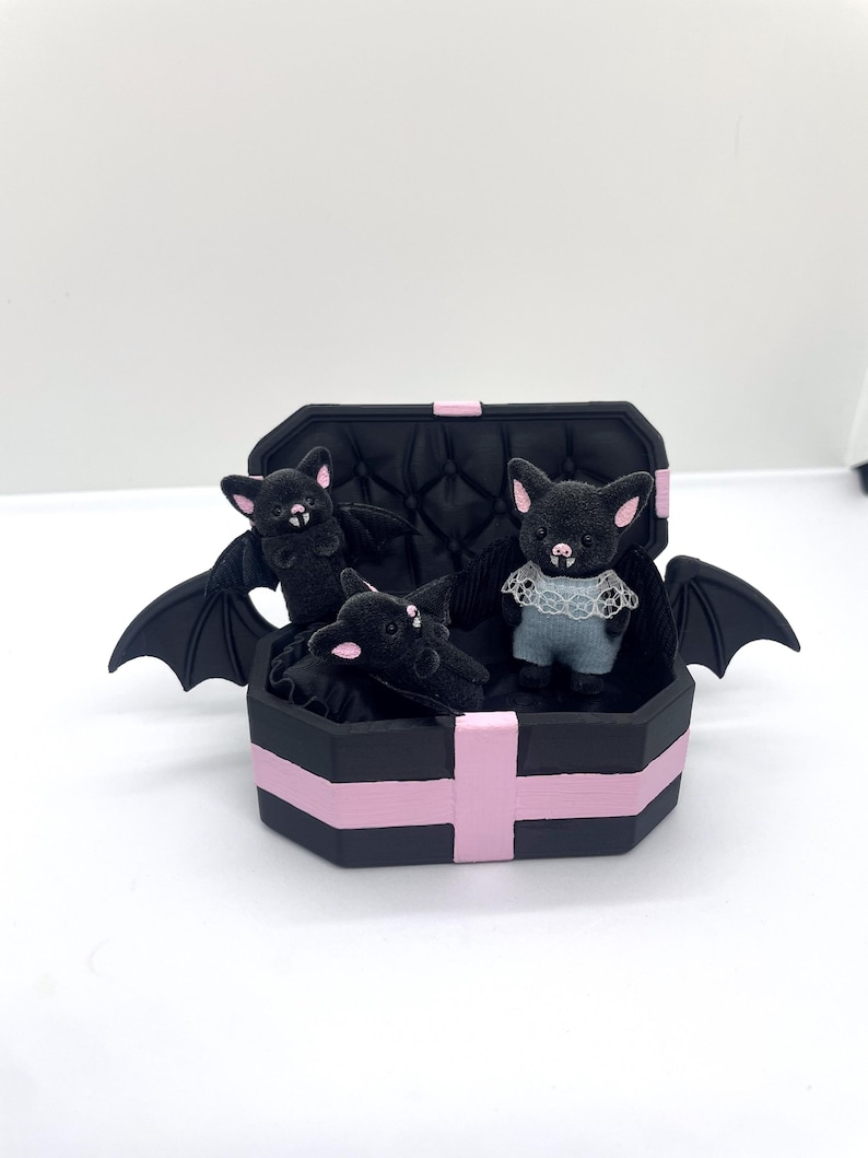 以下が含まれることがあります： ピンクのアクセントが付いた黒い棺型の箱に、3匹のぬいぐるみコウモリが入っています。2匹のコウモリは翼を広げ、3匹目は白いレースの襟が付いた青い服を着ています。コウモリはピンクの耳をしています。