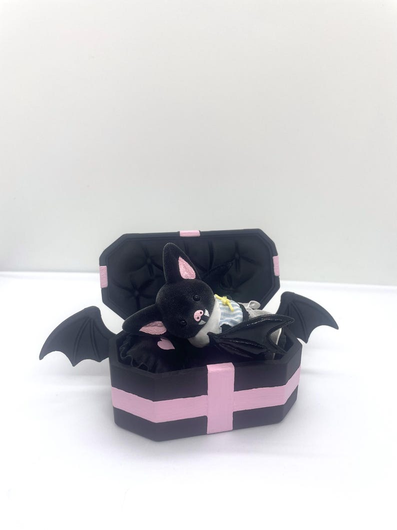 以下が含まれることがあります： ピンクのアクセントとコウモリのぬいぐるみが入った、黒い棺型の箱。 コウモリはピンクの耳をしており、小さな衣装を着ています。 棺には蝶番付きの蓋とコウモリの羽が付いています。