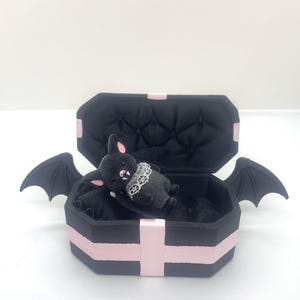 May include: A small black bat figurine with pink inner ears and white fangs, wearing a white lace collar, resting inside a black coffin-shaped box with pink cross accents and black bat wings attached to the sides of the box exterior.