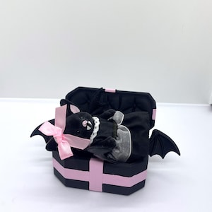 以下が含まれることがあります： ピンクのアクセントとピンクのリボンが付いた、黒い棺型の箱。中には、白い襟と黒いドレスを着た、ぬいぐるみコウモリ人形が入っています。コウモリは黒い翼を持っており、箱の中に横たわっています。箱は開いています。