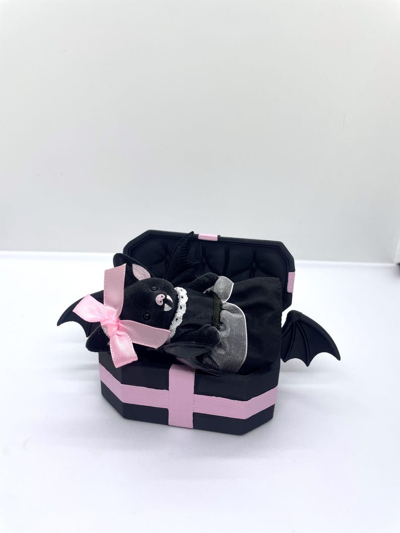 以下が含まれることがあります： ピンクのアクセントとピンクのリボンが付いた、黒い棺型の箱。中には、白い襟と黒いドレスを着た、ぬいぐるみコウモリ人形が入っています。コウモリは黒い翼を持っており、箱の中に横たわっています。箱は開いています。