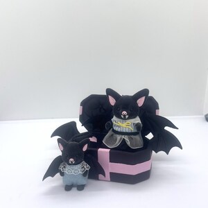 以下が含まれることがあります： ピンクのアクセントが付いた2つの黒いコウモリの置物。1つは立っていて、もう1つは黒とピンクの箱の上にいます。コウモリは大きな翼を持ち、レースと青いディテールのある衣装を着ています。箱はコウモリの羽のデザインです。