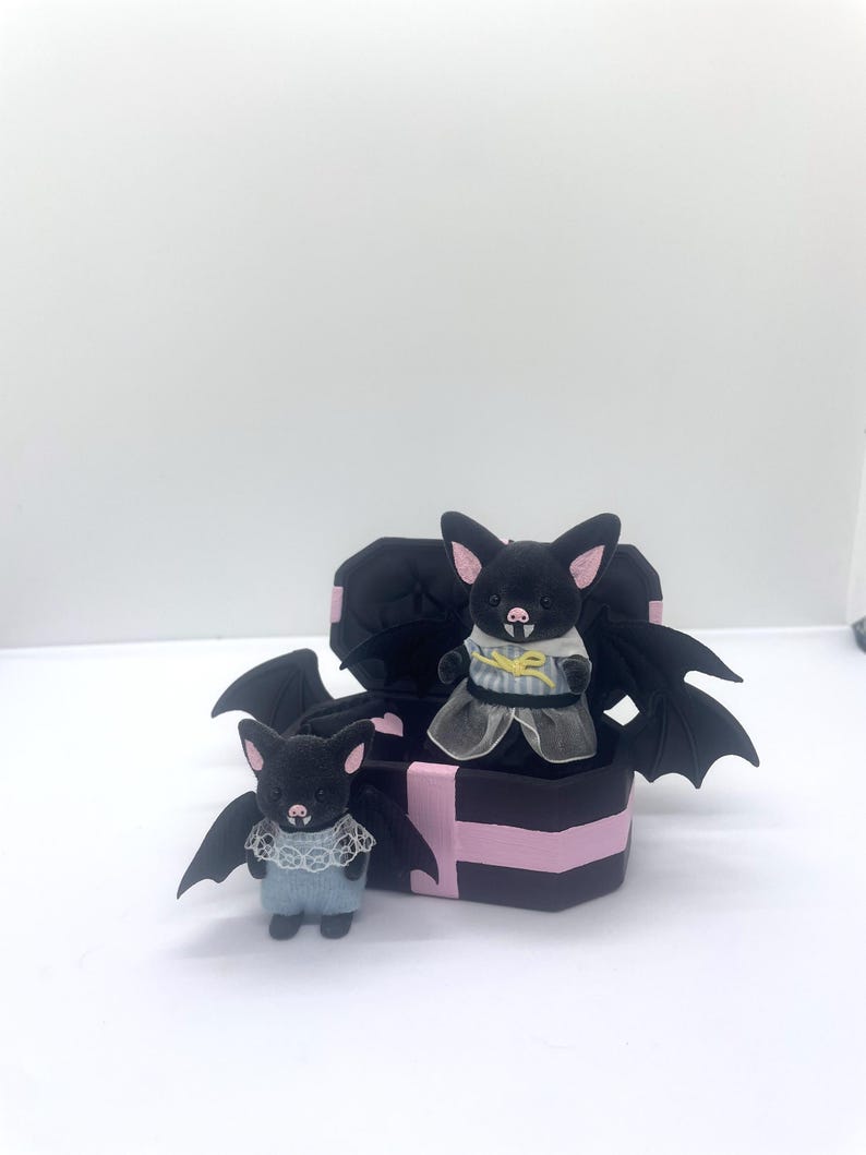 以下が含まれることがあります： ピンクのアクセントが付いた2つの黒いコウモリの置物。1つは立っていて、もう1つは黒とピンクの箱の上にいます。コウモリは大きな翼を持ち、レースと青いディテールのある衣装を着ています。箱はコウモリの羽のデザインです。