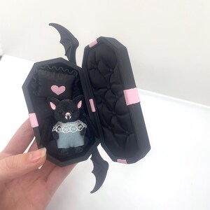 以下が含まれることがあります： ピンクのアクセントが付いた黒い棺型の箱で、開くとぬいぐるみコウモリの人形が現れます。 コウモリは青い衣装とレースの襟を着ており、ピンクのハートが付いた枕の上にいます。 棺にはコウモリの羽のディテールがあります。